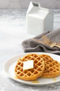 A plate with two waffles topped with a pat of butter and syrup, with a milk jug in the background and a fork on a cloth napkin to the side.