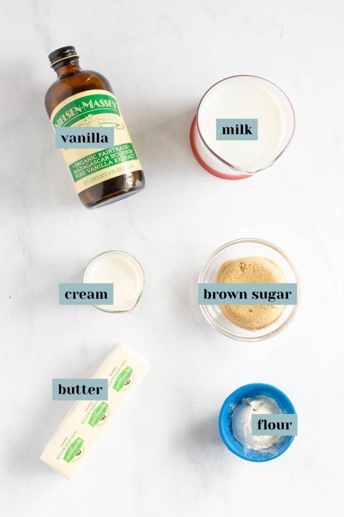Flat lay of labeled ingredients including vanilla extract, milk, cream, brown sugar, butter, and flour on a light surface.