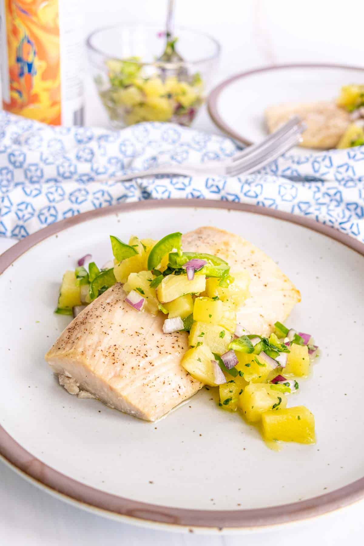 Mahi mahi topped with pineapple salsa served on a white plate with a blue patterned tablecloth, and a side salad in the background.