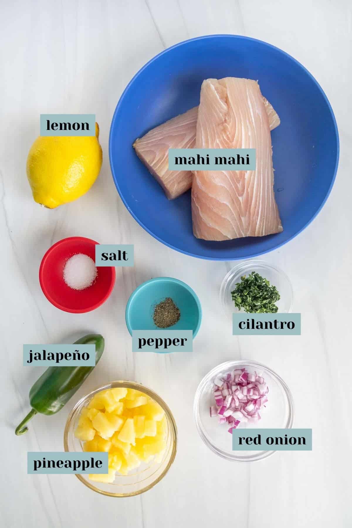 Ingredients for cooking spread on a table, including mahi mahi fillets in a blue bowl, and small bowls with lemon, salt, jalapeño, pineapple, cilantro, and red onion.