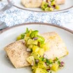 White fish fillets topped with a pineapple and red onion salsa, served on a beige plate with a decorative blue and white napkin in the background.