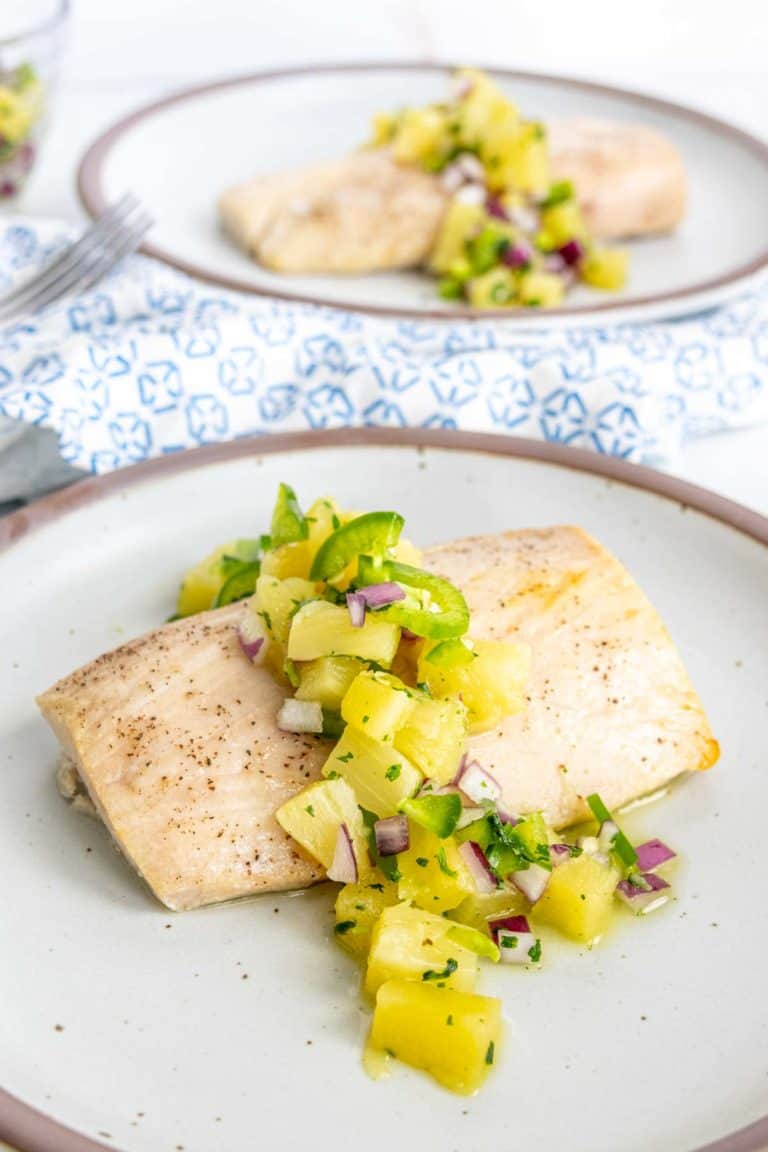 White fish fillets topped with a pineapple and red onion salsa, served on a beige plate with a decorative blue and white napkin in the background.