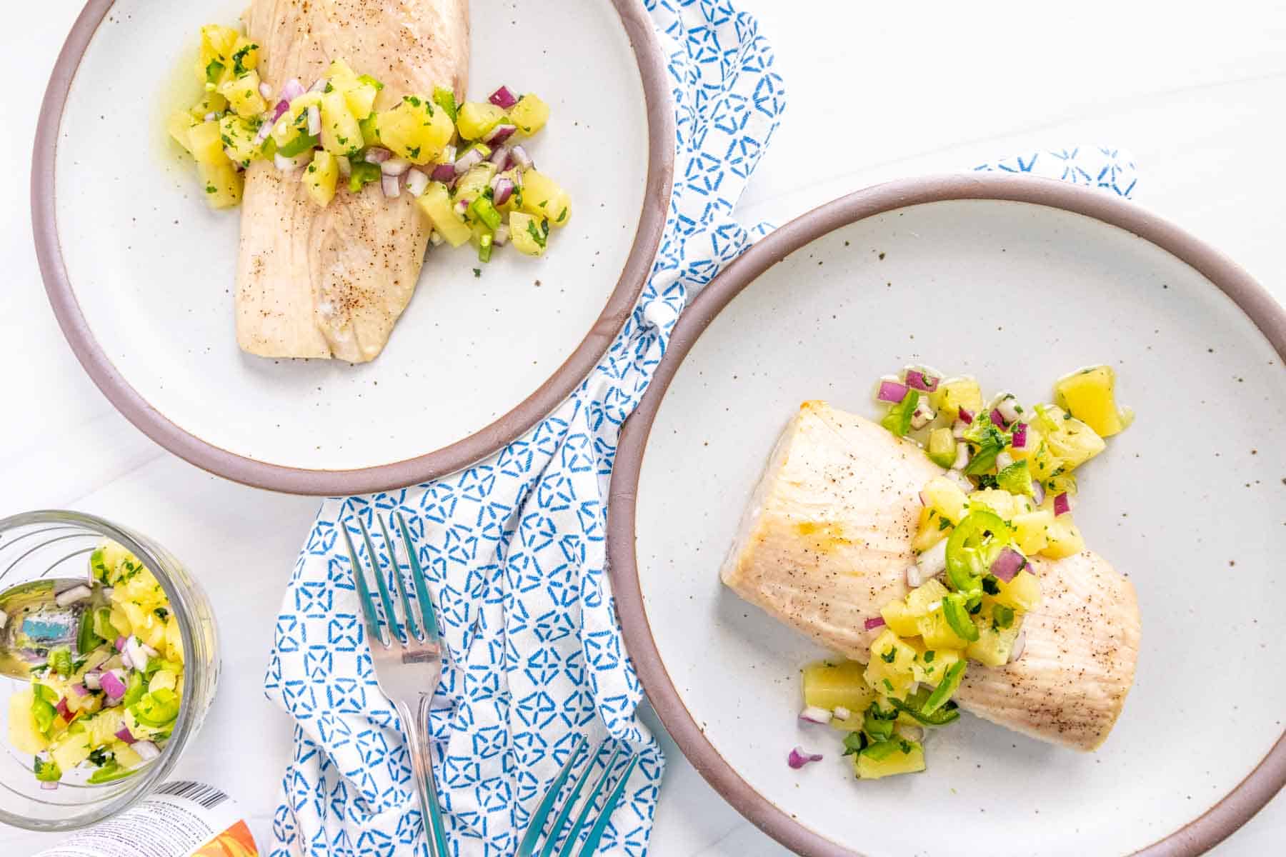 Two plates with fish fillets topped with pineapple and red onion salsa, set on a white table with a patterned napkin between them.