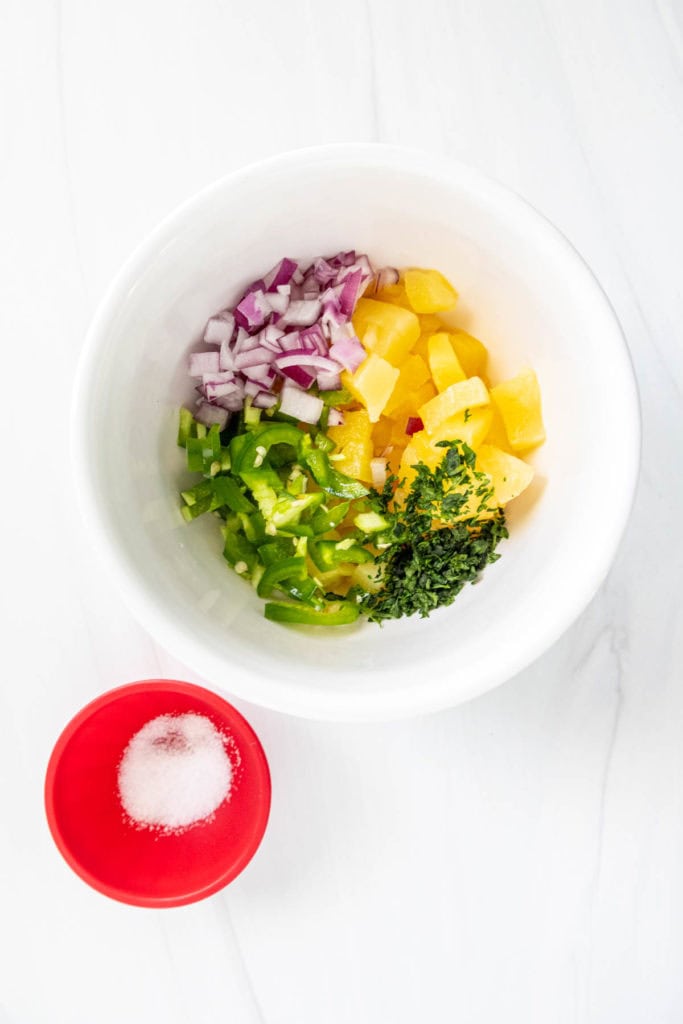 A white bowl containing chopped peppers, onions, pineapple, and herbs, with a small red bowl of salt beside it.