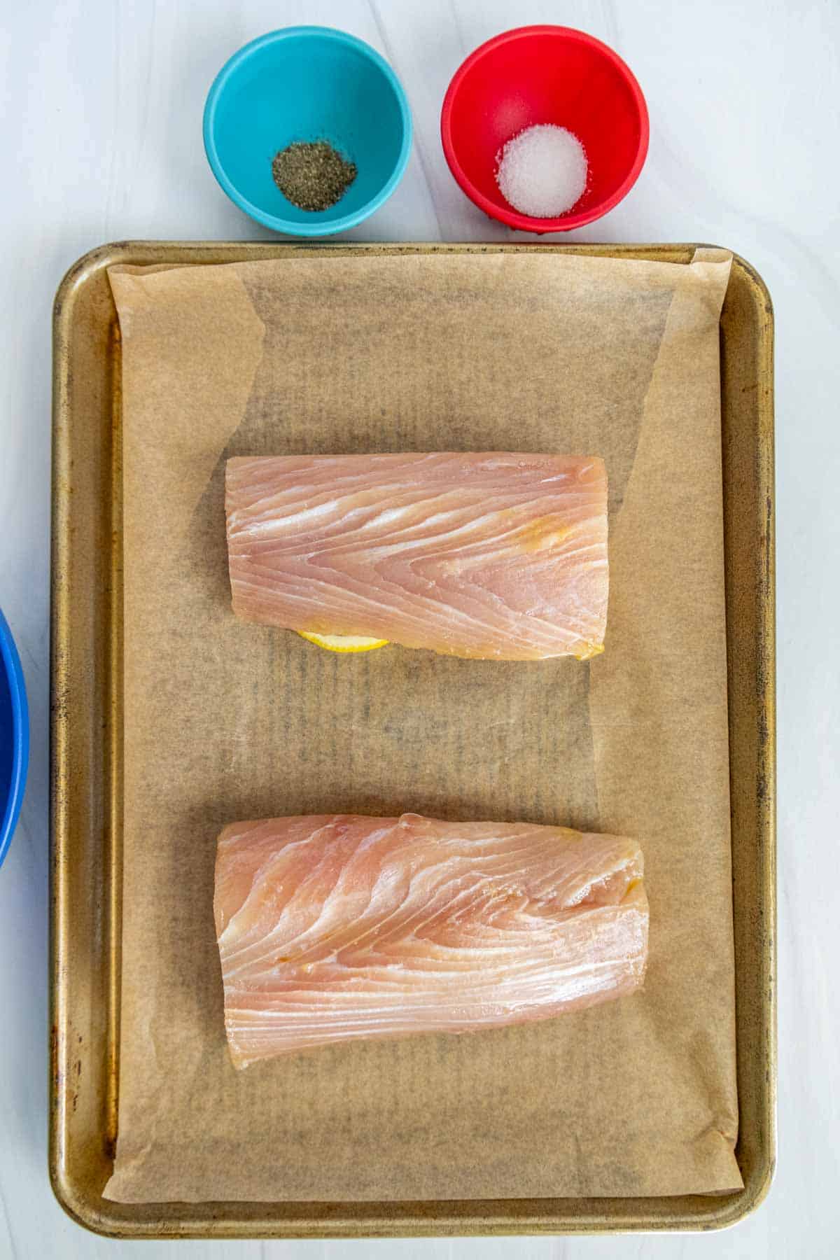 Two raw fish fillets on a parchment-lined baking tray, with small bowls containing salt, pepper, and other spices beside them.