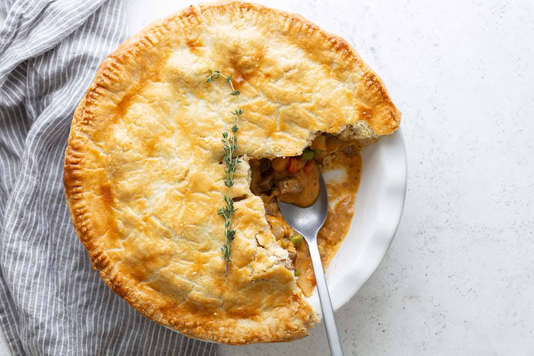 A homemade beef pot pie with a flaky crust, partially sliced to show its creamy filling of beef and vegetables, garnished with fresh thyme.