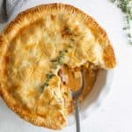 A golden beef pot pie with a slice removed, revealing its creamy filling, garnished with thyme on a white plate with a spoon.