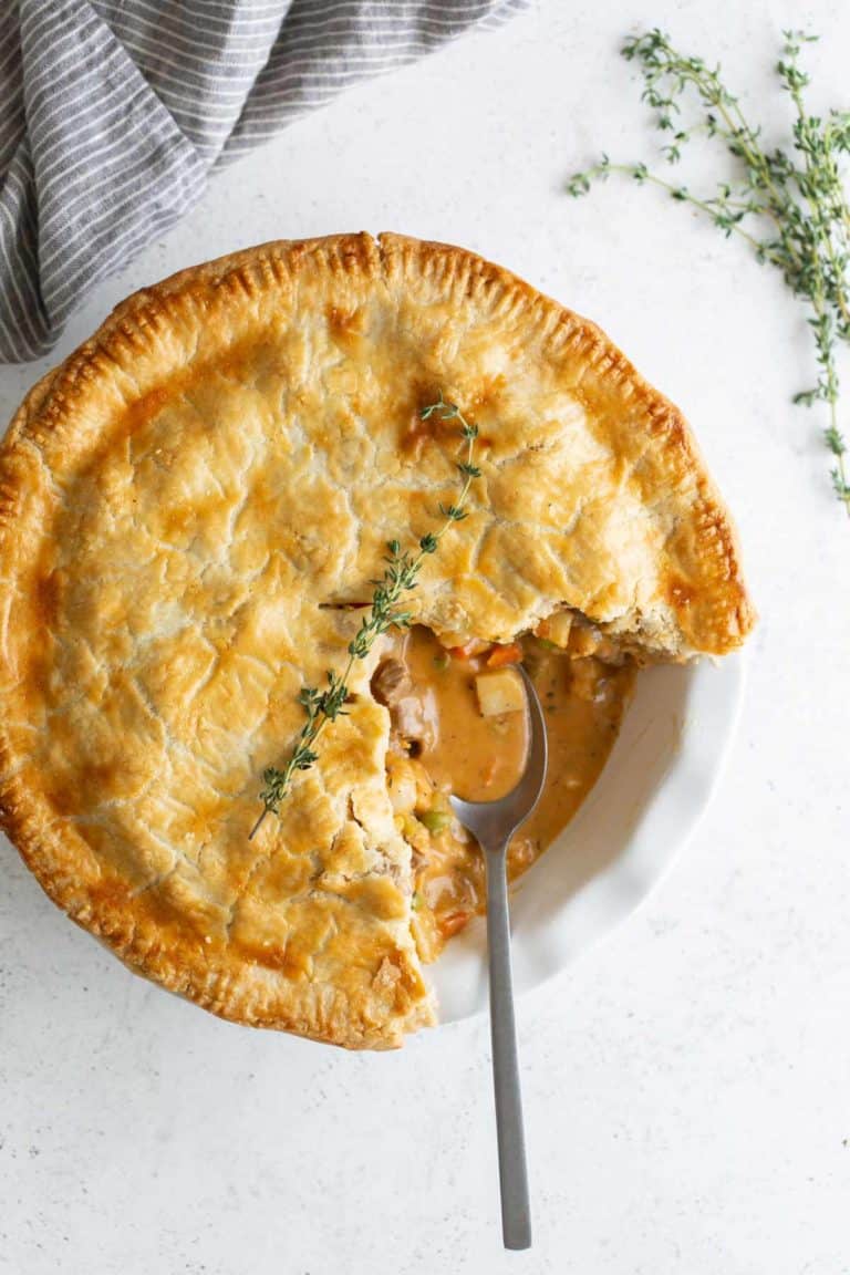 A golden beef pot pie with a slice removed, revealing its creamy filling, garnished with thyme on a white plate with a spoon.