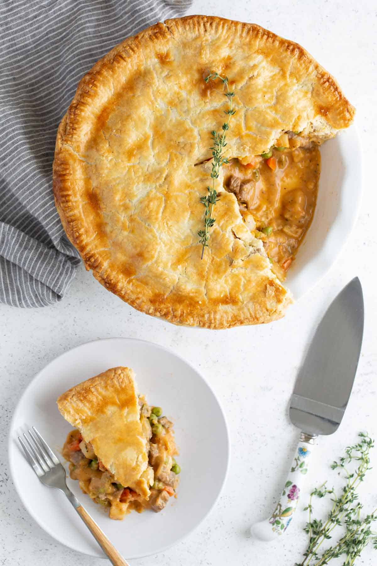 Beef pot pie with a slice served on a white plate, accompanied by a knife and fresh thyme sprigs on a marble surface.