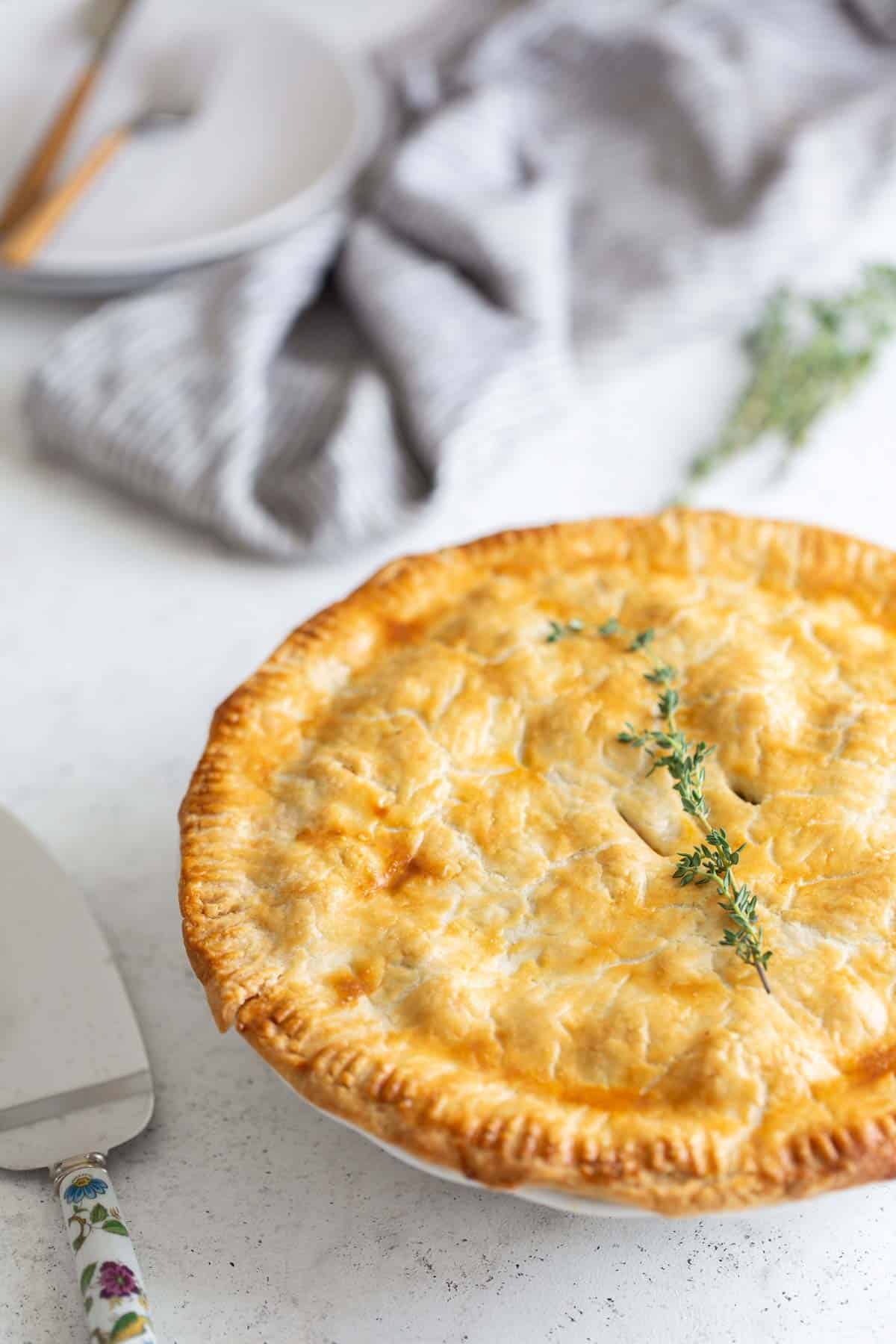 Golden baked pie with a flaky crust and a thyme garnish, served on a white dish with cutlery and a napkin on a light background.