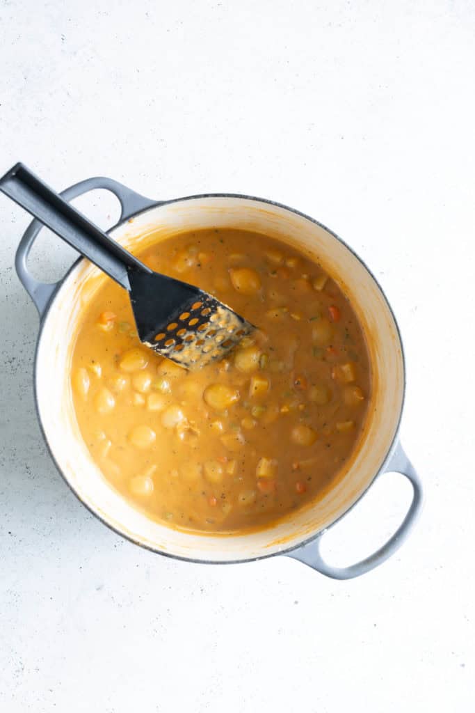 A pot of creamy vegetable soup with sliced carrots and peas, a ladle inside, on a light textured surface.