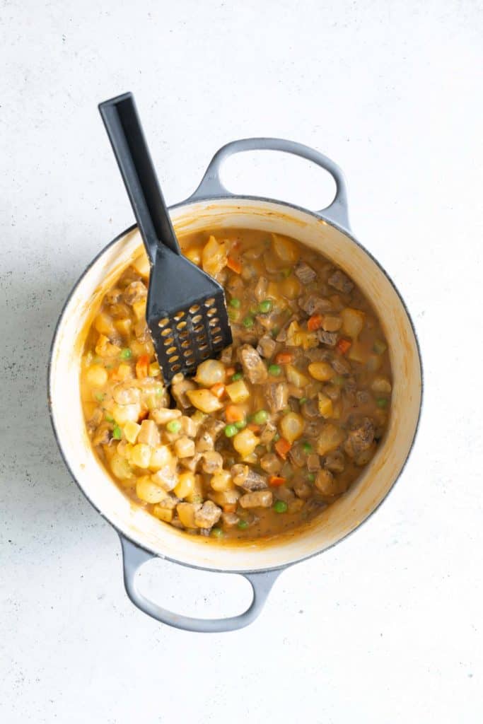 Creamy beef and vegetable stew in a light blue pot with a black ladle, viewed from above on a textured white surface.