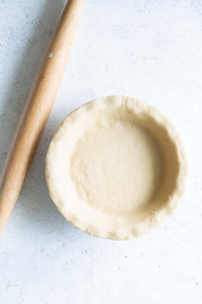 Unbaked pie crust in a dish next to a wooden rolling pin on a light-colored surface.
