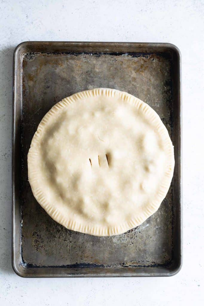 Unbaked pie with a crimped edge and vent slits on a worn baking sheet, top view.