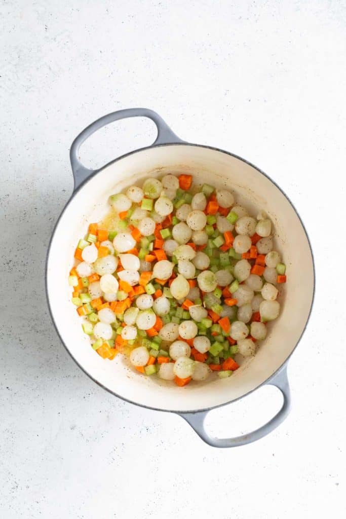 A white pot containing onions, carrots, and celery, chopped and prepped for cooking, viewed from above on a light surface.