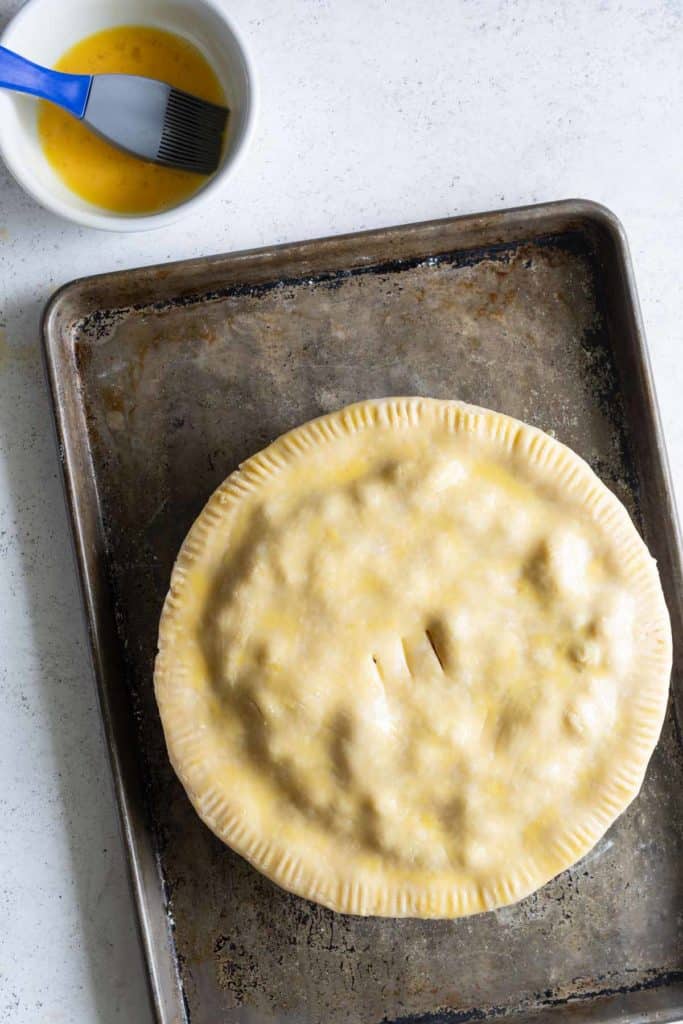 An uncooked pie with a vented crust on a baking sheet, next to a brush and a bowl of egg wash on a white surface.