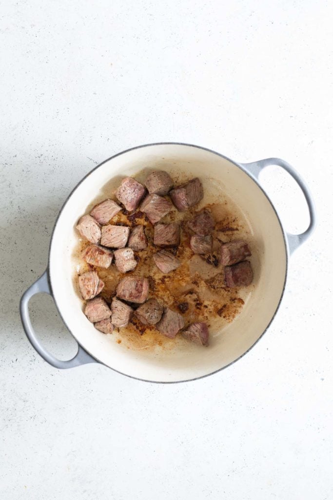 Seared cubes of beef in a white dutch oven on a light surface.