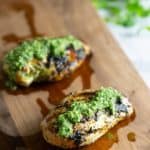 Two grilled chicken breasts topped with chimichurri sauce on a wooden cutting board, with some herbs in the background.