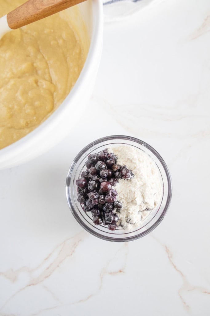A glass cup filled with flour and blueberries sits on a marble countertop next to a mixing bowl containing batter.