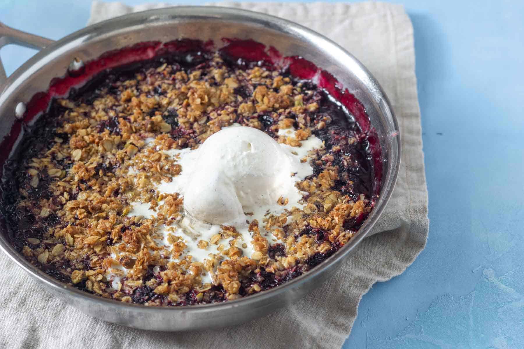 A skillet containing a berry crumble topped with a scoop of vanilla ice cream sits on a beige cloth.