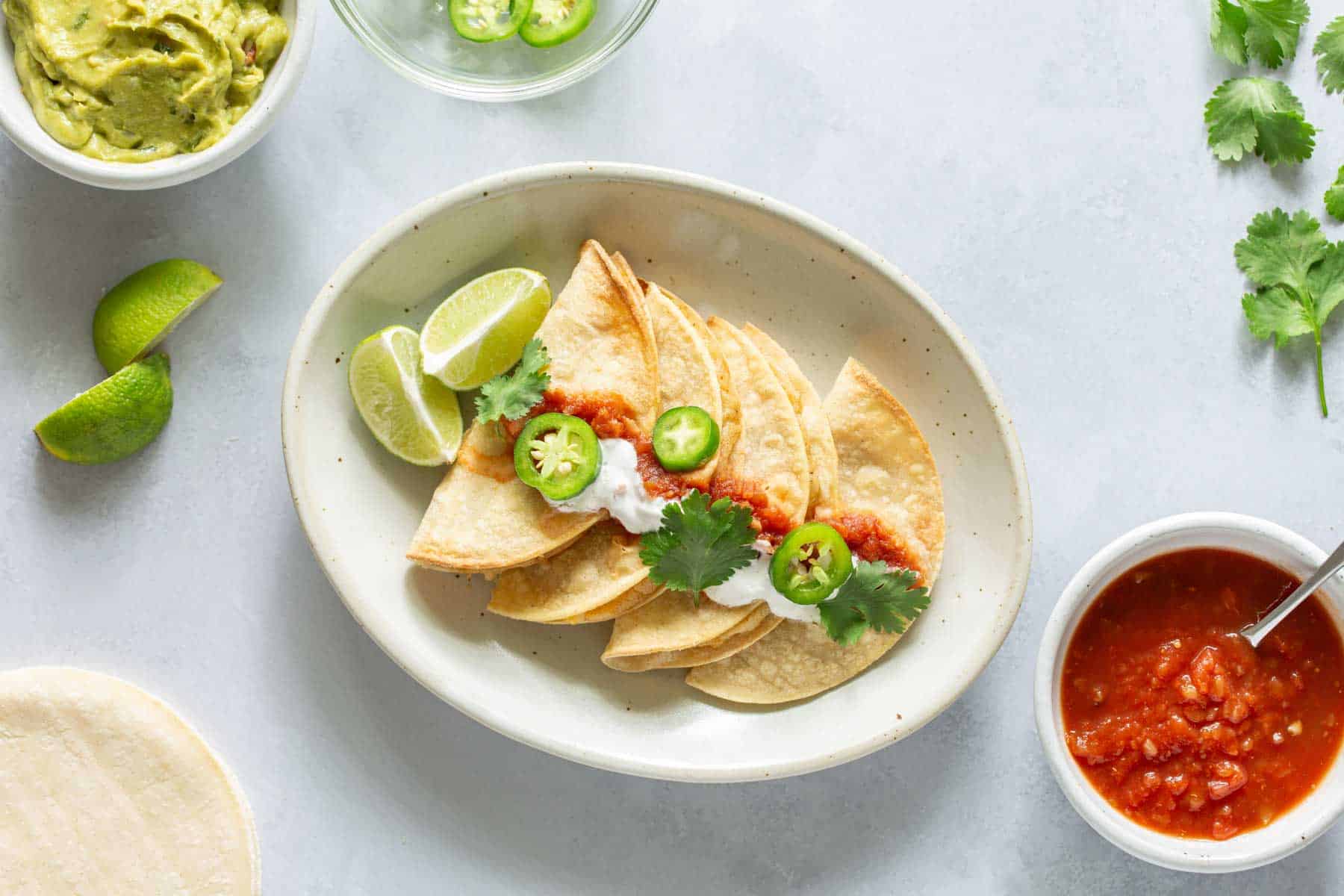 A white plate with four folded tacos topped with sour cream, salsa, jalapeño slices, and cilantro. It is garnished with lime wedges. Surrounding the plate are bowls of salsa, guacamole, tortillas, and lime.