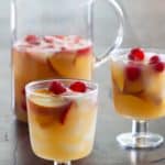 A pitcher and two glasses filled with sangria, garnished with floating raspberries and peach slices on a wooden table.