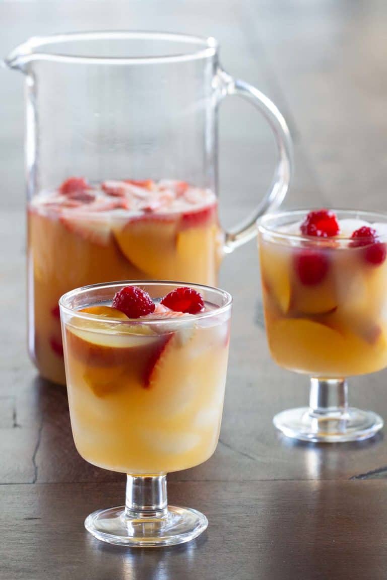 A pitcher and two glasses filled with sangria, garnished with floating raspberries and peach slices on a wooden table.