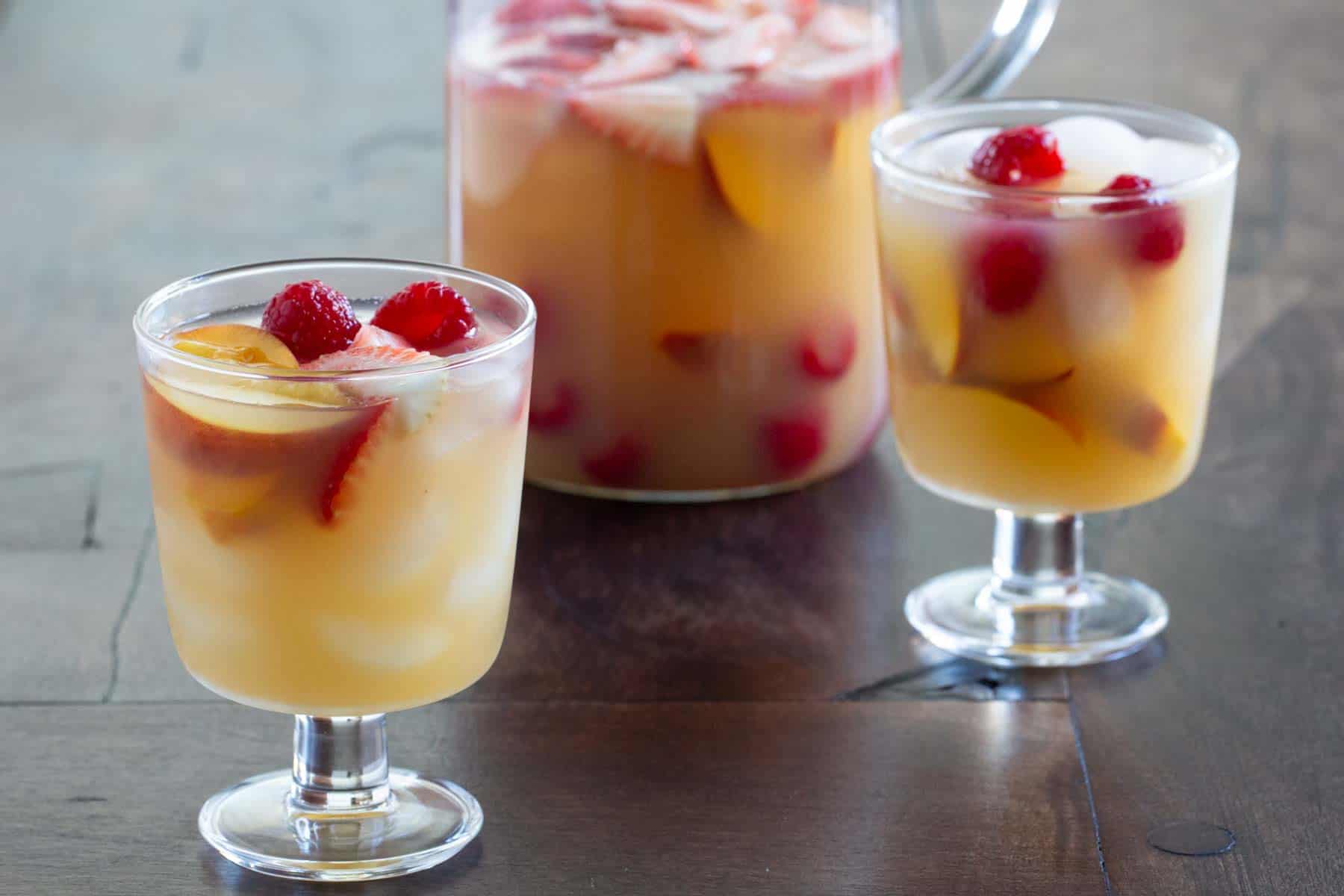 Two glasses and a pitcher of fruit sangria on a wooden table, containing sliced strawberries, peaches, and cherries.