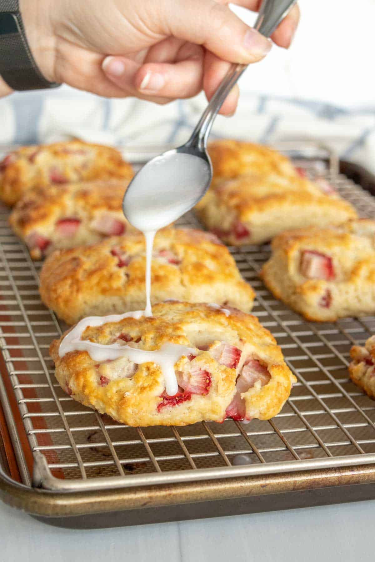 A hand drizzling icing on fresh strawberry biscuits cooling on a wire rack.
