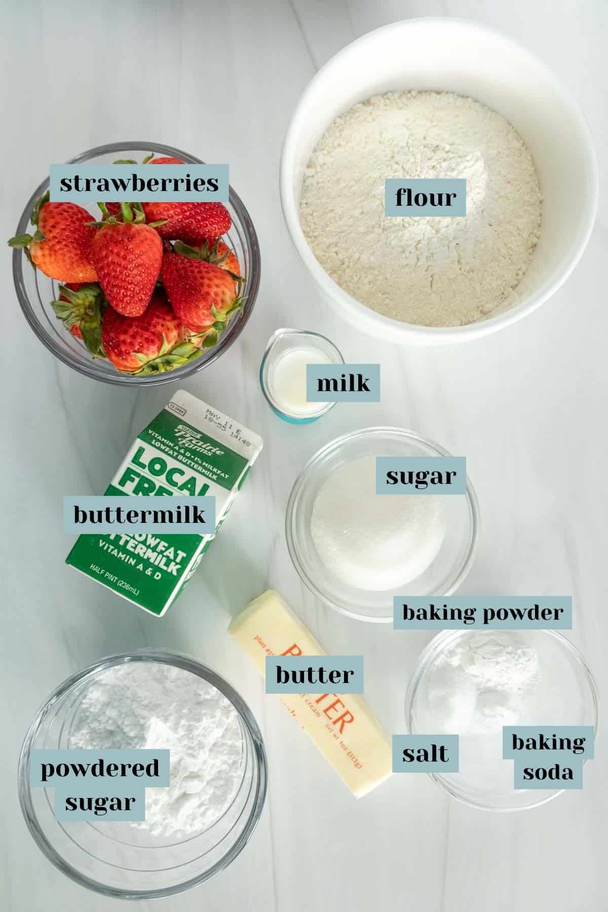 Ingredients for baking laid out on a white surface, including strawberries, flour, milk, sugar, buttermilk, butter, baking powder, baking soda, powdered sugar, and salt, labeled for identification.