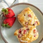 Two strawberry biscuits with icing on a beige plate, accompanied by fresh strawberries, on a white table with a light blue-striped napkin.