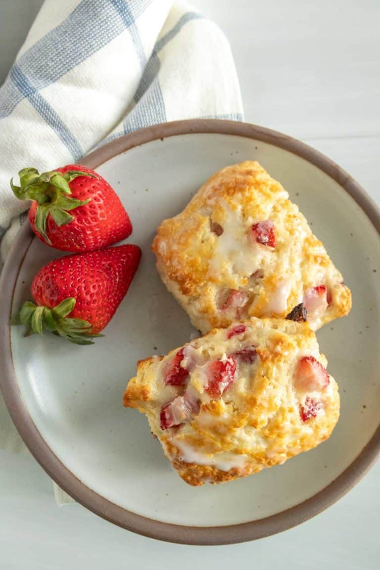 Two strawberry biscuits with icing on a beige plate, accompanied by fresh strawberries, on a white table with a light blue-striped napkin.