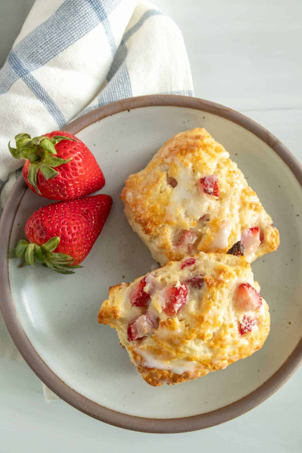 Two strawberry biscuits with icing on a beige plate, accompanied by fresh strawberries, on a white table with a light blue-striped napkin.