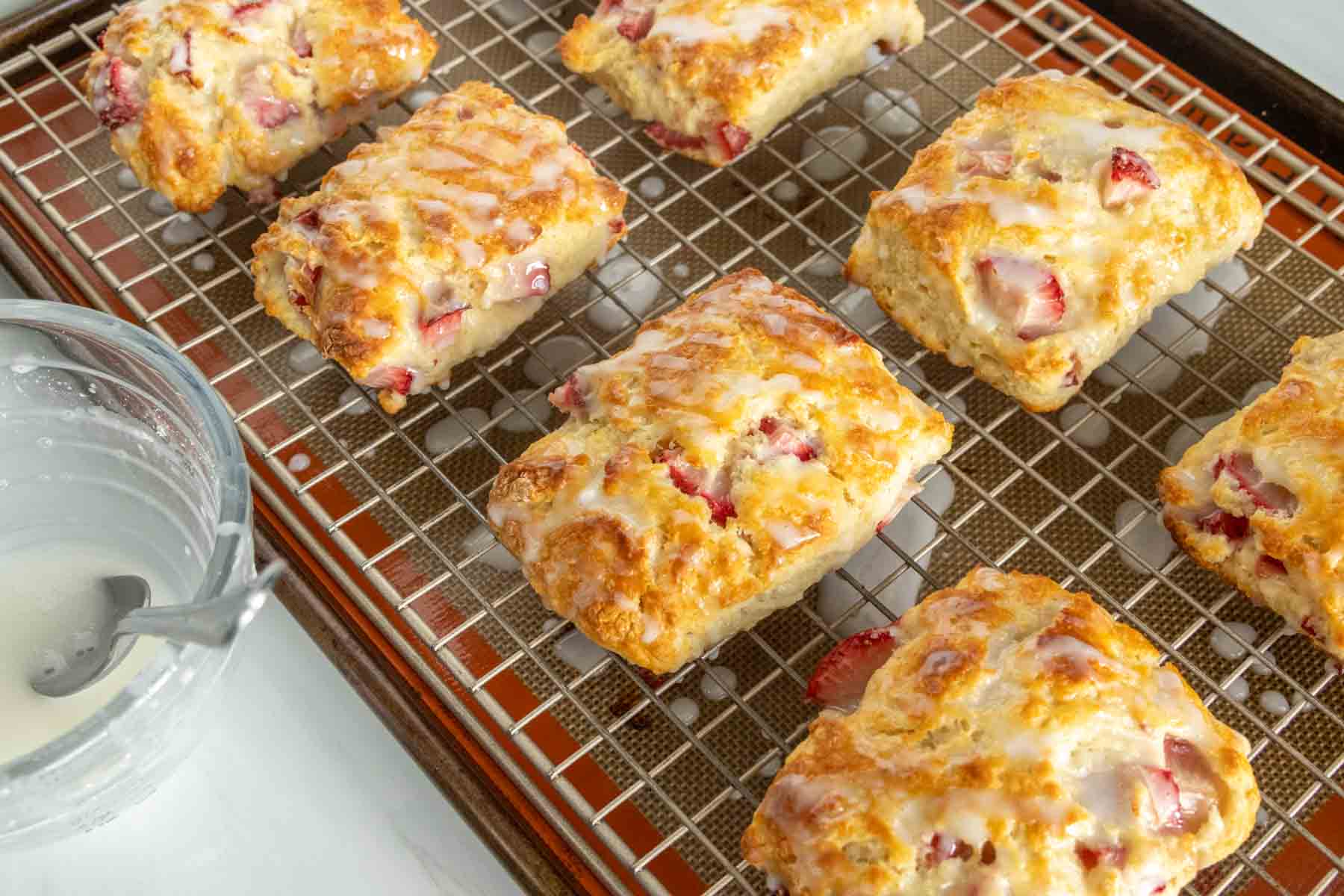 Freshly baked strawberry biscuits with glaze on a cooling rack beside a glass jar of glaze.