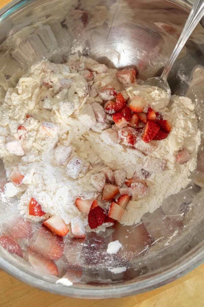 A stainless steel mixing bowl containing flour and sliced strawberries, with a spoon partially visible, on a wooden surface.