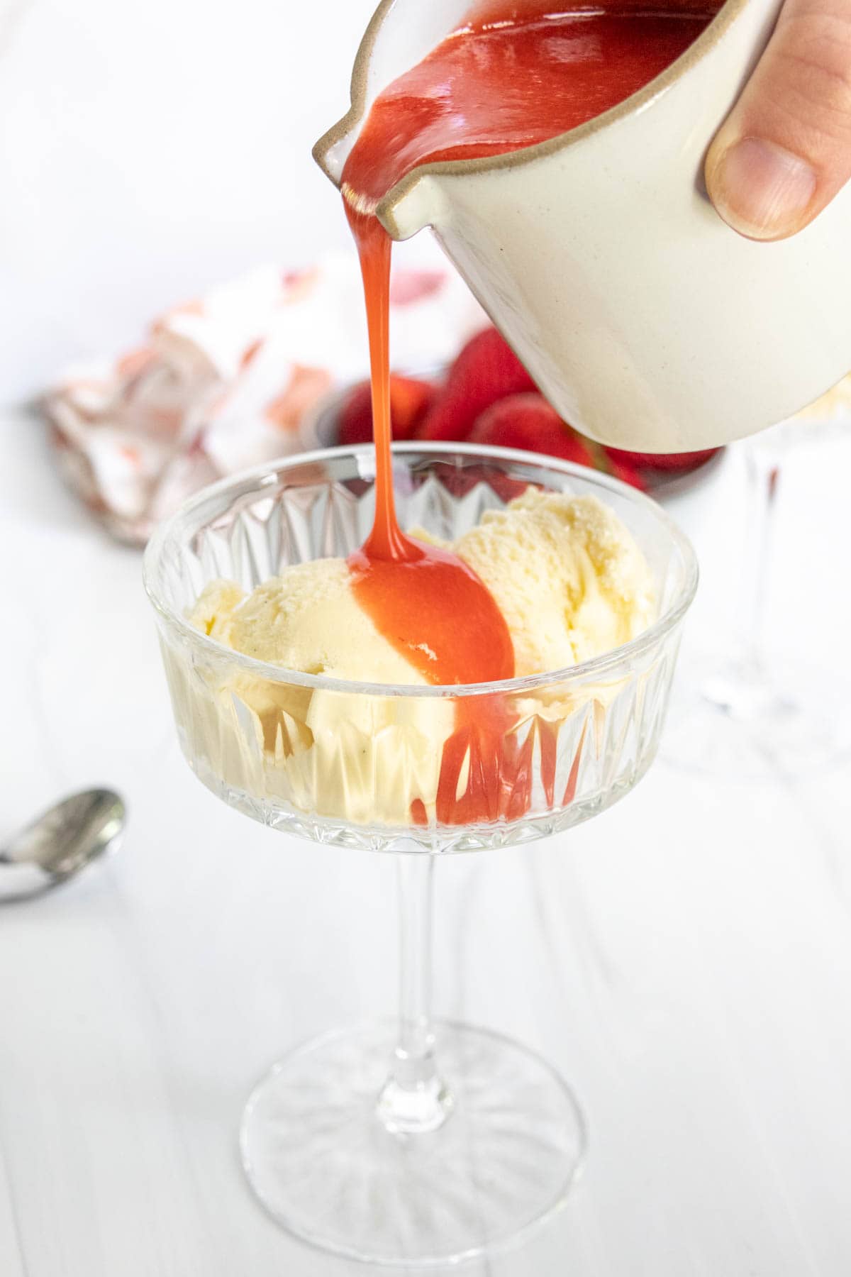 A person pouring strawberry syrup over vanilla ice cream in a glass bowl on a white table.