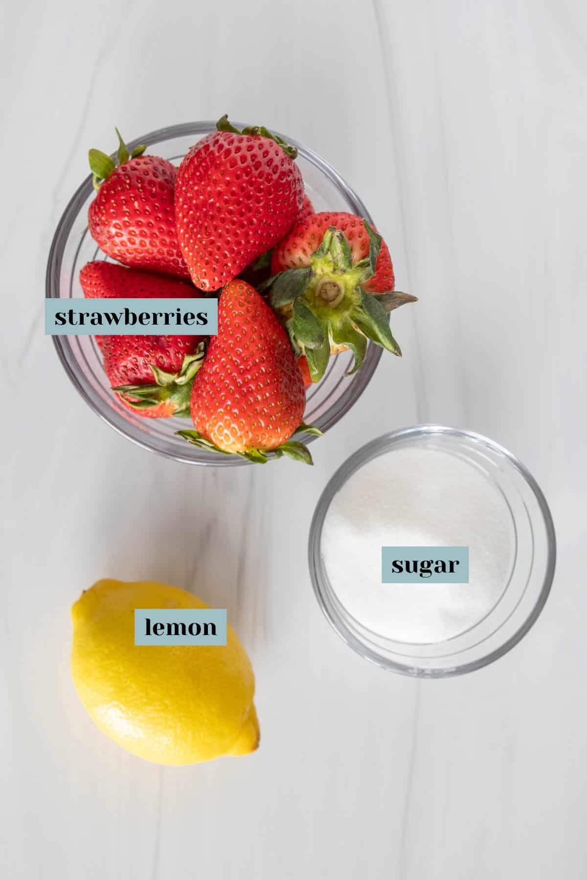 A top-down view of ingredients for a recipe on a white surface: a bowl of strawberries, a lemon to the side, and a smaller bowl of sugar, each labeled.