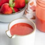A ceramic jug filled with strawberry sauce, with whole strawberries in a bowl and a filled glass jar in the background.