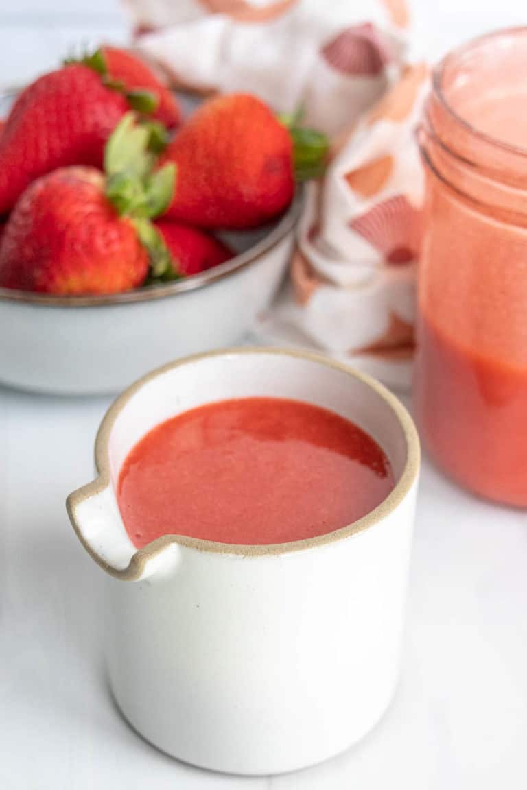 A ceramic jug filled with strawberry sauce, with whole strawberries in a bowl and a filled glass jar in the background.
