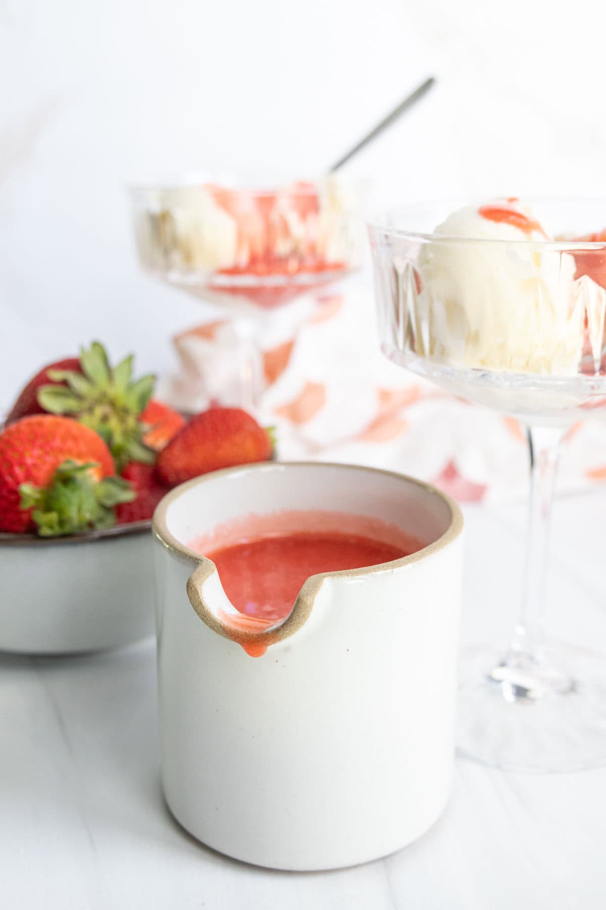 A ceramic pot of strawberry sauce with fresh strawberries, a bowl of whipped cream, and dessert glasses in the background on a white surface.