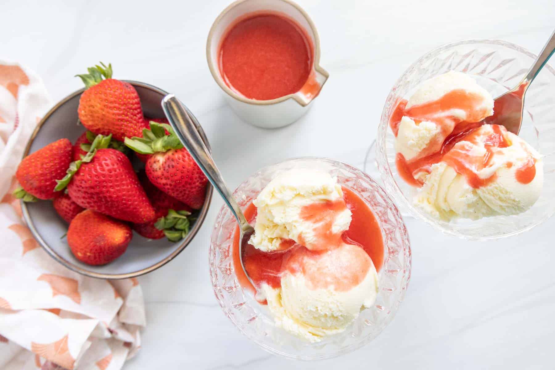 Two bowls of vanilla ice cream topped with strawberry sauce, accompanied by a bowl of fresh strawberries and a small pitcher of extra sauce on a marble countertop.