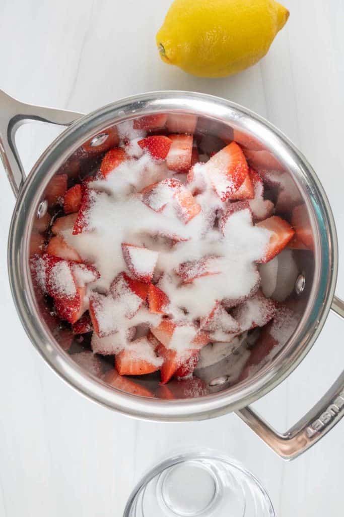 Top view of a stainless steel pot containing sliced strawberries and sugar with a whole lemon on the side, on a white surface.