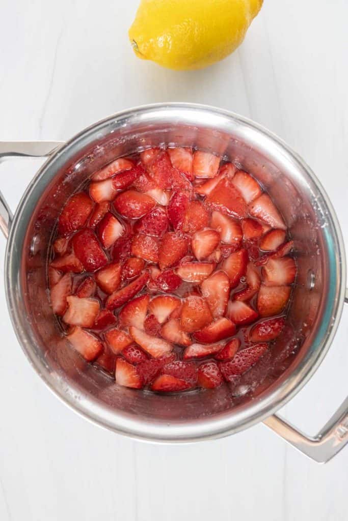 A saucepan filled with sliced strawberries in syrup, placed on a white wooden surface next to a whole lemon.