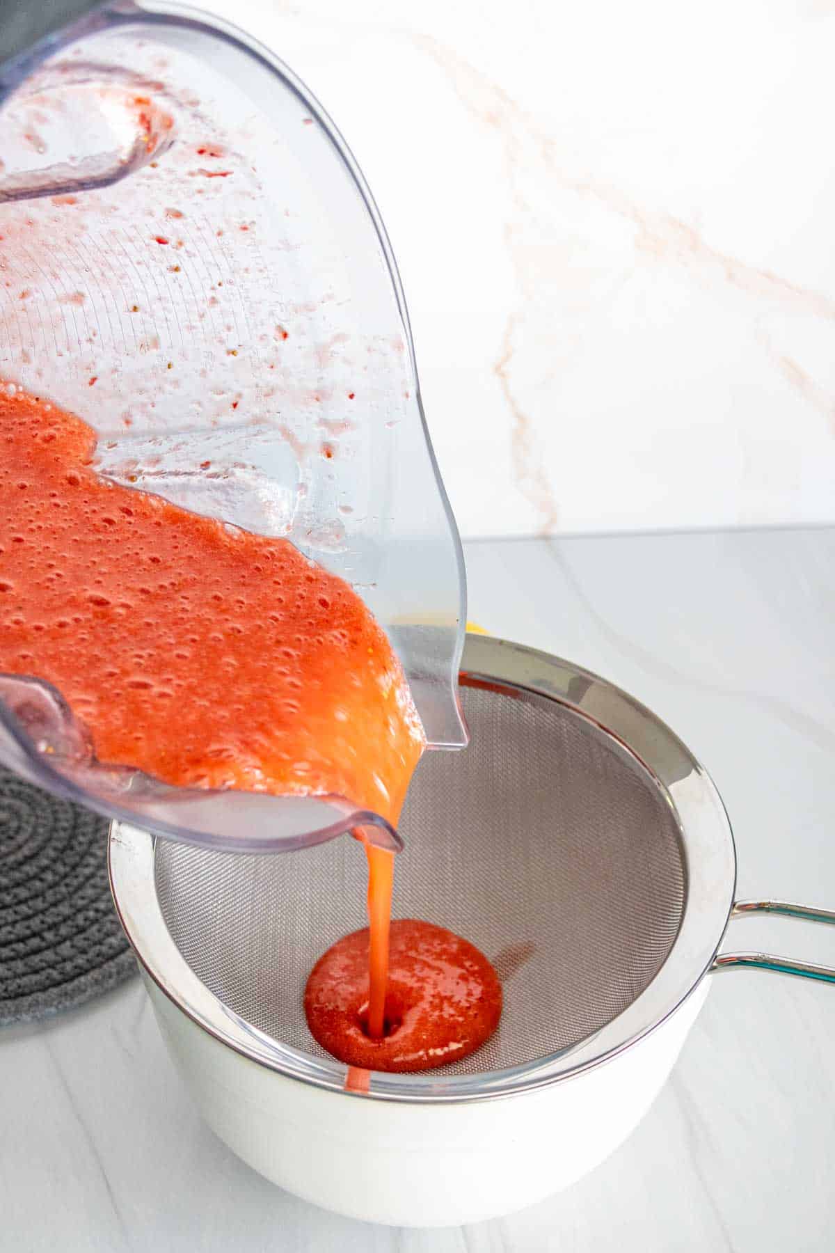 Pouring freshly blended strawberry puree through a fine mesh sieve into a white bowl on a marble countertop.