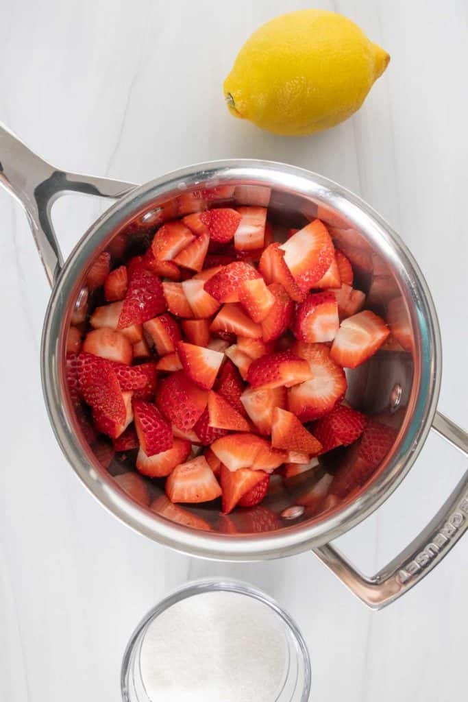 Top view of a stainless steel pot filled with chopped strawberries, alongside a whole lemon and a glass lid, on a white surface.
