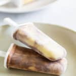 Two swirl-patterned homemade popsicles lie on a beige plate, with more popsicles blurred in the background.
