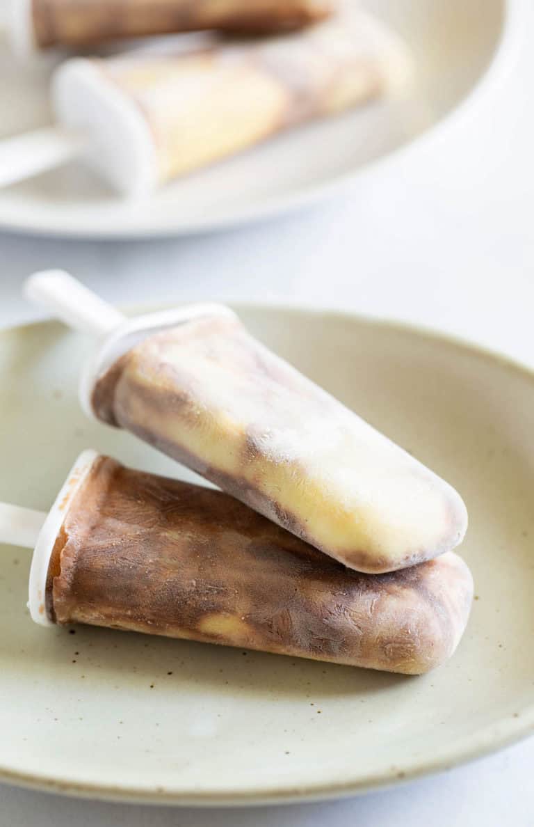 Two swirl-patterned homemade popsicles lie on a beige plate, with more popsicles blurred in the background.