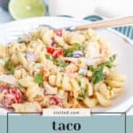 A plate of taco pasta salad with chicken, garnished with cilantro and red onion, sits in front of a baking dish; a halved lime and creamy avocado are in the background.