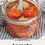 A glass jar of tomato confit, brimming with cherry tomatoes and garlic cloves steeped in oil, with a spoon nestled inside.