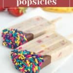 Two delicious banana popsicles, partially dipped in chocolate and colorful sprinkles, are displayed on a white surface, with fresh strawberries and bananas in the background.